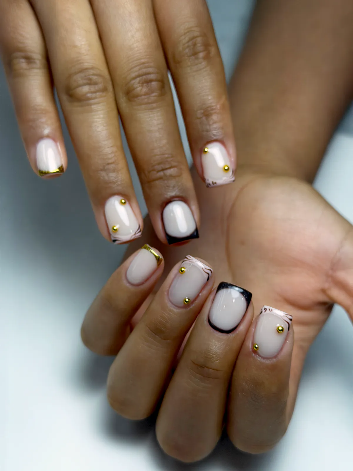 Short square milky-nude nails with black and gold chrome micro-French tips and gold studs, by BeautyFusionClau in Sarasota, Florida